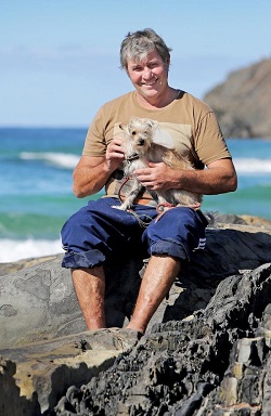 A photo of Terry sitting on a rock on the beach with a dog sitting on his lap